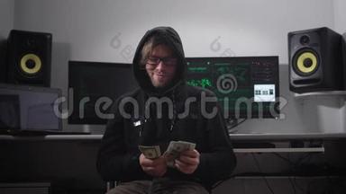 一个穿着夹克衫，戴着引擎盖的人在电脑室。 戴眼镜、手<strong>拿钱</strong>的黑客坐在椅子上。 一个男人